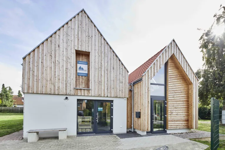 Zwei ineinander übergehende Häuser. Ein Haus hat ein weißes Erdgeschoß und Holzverkleidung am Giebel, das andere Haus ist vollständig mit Holz bekleidet